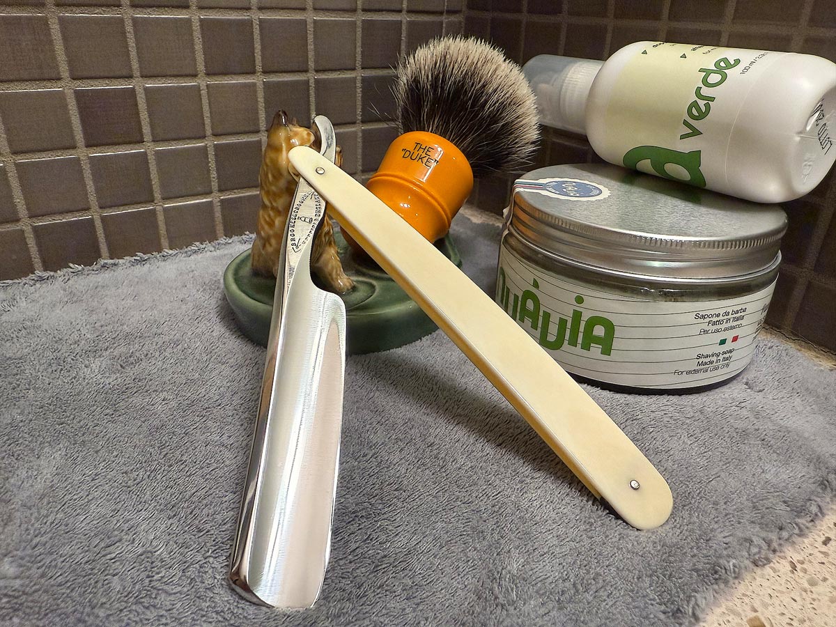 On a gray microfiber towel a straight razor with blade open about 45 degrees leans next to a bottle of aftershave and a jar of shave soap. A shave brush rests bristles-up in a green Wade ceramic pipe rest that features a brown Cairn Terrier.