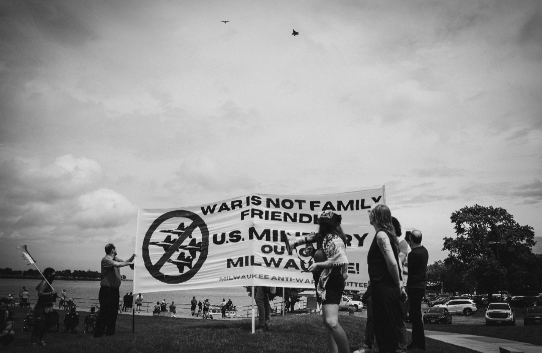Protesters hold a banner as a fighter jet flies above.
