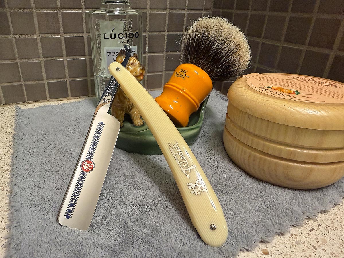 On a gray microfiber towel a straight razor with blade open about 45 degrees leans next to a bottle of aftershave and a jar of shave soap. A shave brush rests bristles-up in a green Wade ceramic pipe rest that features a brown Cairn Terrier.