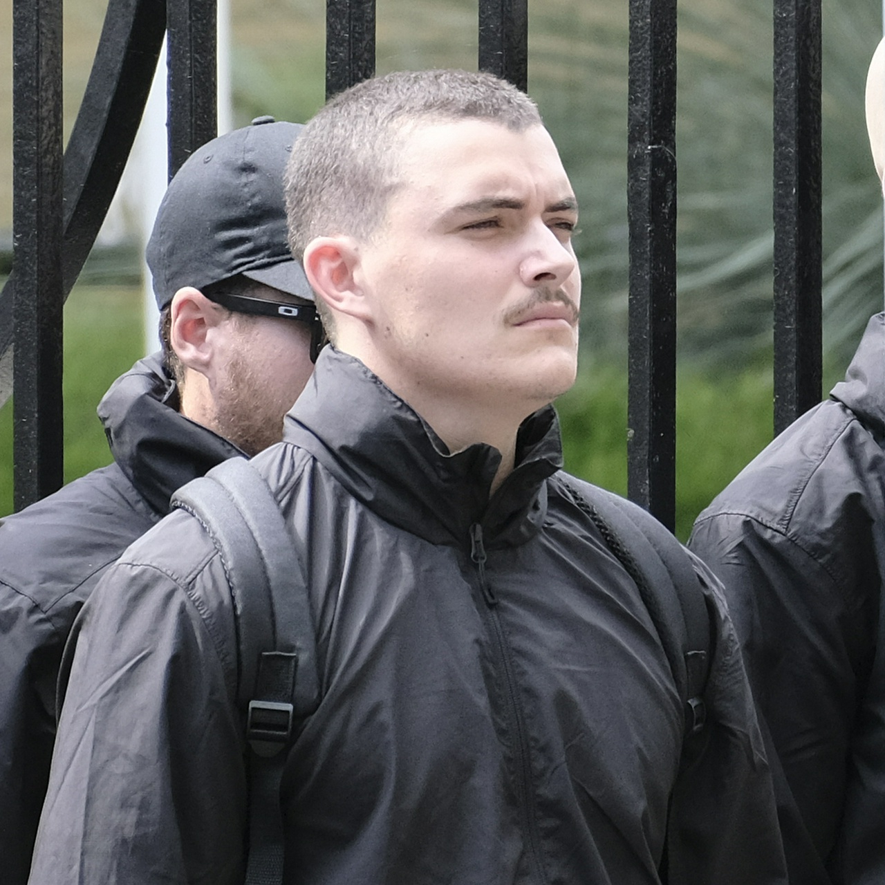 A stocky white man with very short brown hair, a bulging temple, a thin moustache and a squint