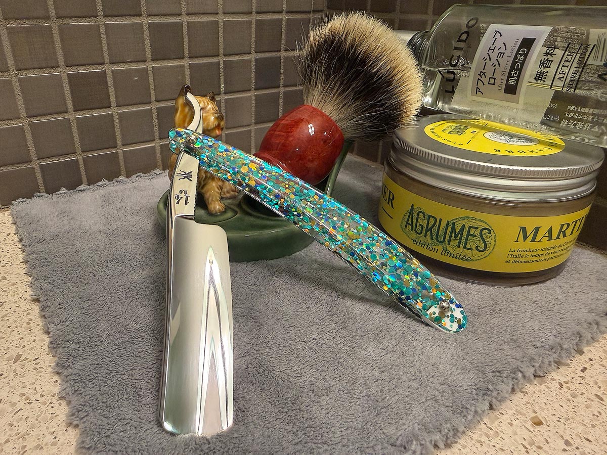 On a gray microfiber towel a straight razor with blade open about 45 degrees leans next to a bottle of aftershave and a jar of shave soap. A shave brush rests bristles-up in a green Wade ceramic pipe rest that features a brown Cairn Terrier.