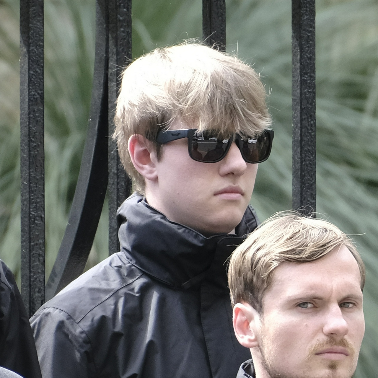 A young guy with a large nose and round chin whose dusty blonde hair falls messily over his thick black sunglasses