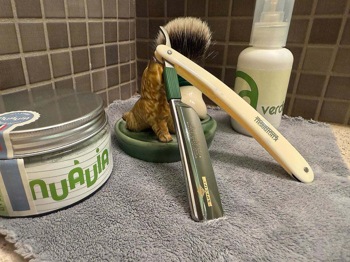 On a gray microfiber towel a straight razor with blade open about 45 degrees leans next to a bottle of aftershave and a jar of shave soap. A shave brush rests bristles-up in a green Wade ceramic pipe rest that features a brown Cairn Terrier.