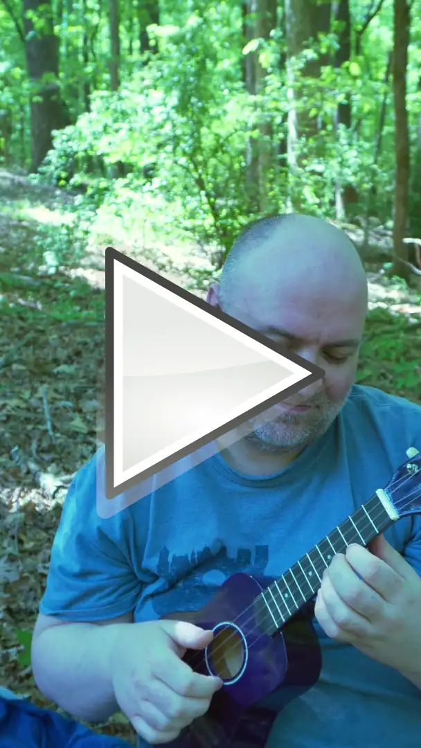 White enby with greying short hair and stubble sitting in front of some trees and holding a purple ukulele.