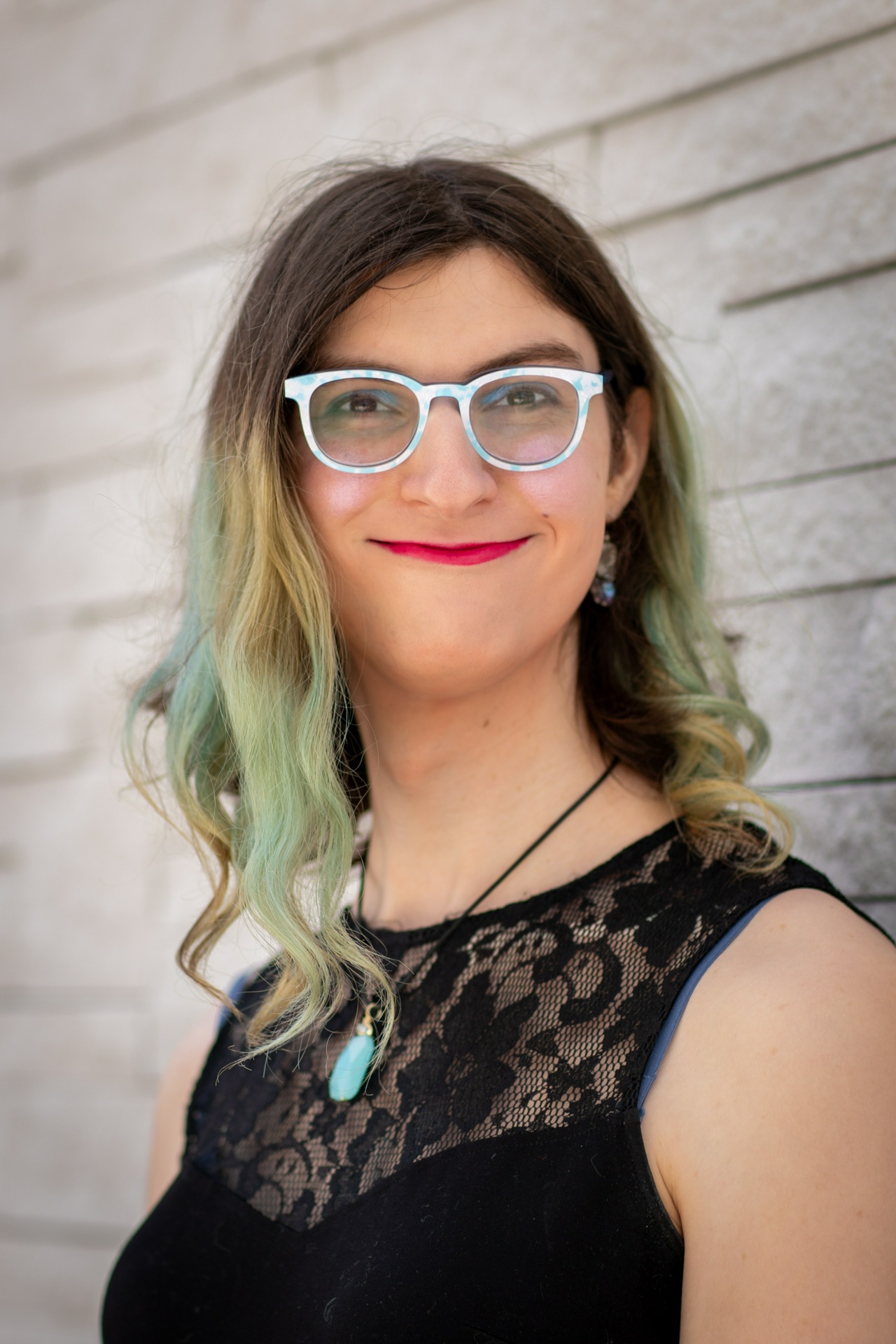 A white girl with brown and blue-green hair down to her shoulders.  She's wearing glasses and has sparkly pink cheeks, along with blue eyeshadow.  Her glasses have a blue and white cloudy sky pattern on them.  She's wearing a black, sleeveless dress, just visible as the picture is focused on her head.  She has a blue pendant on.