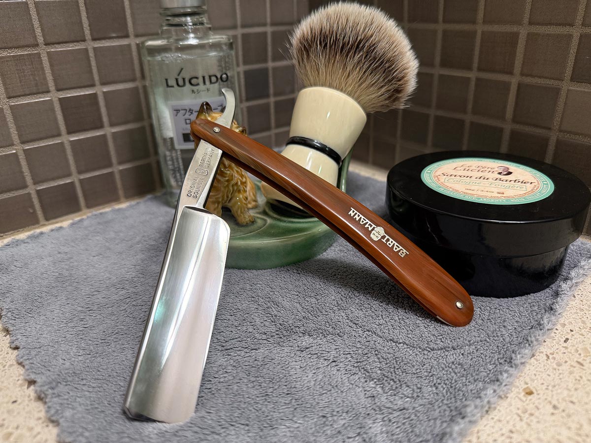 On a gray microfiber towel a straight razor with blade open about 45 degrees leans next to a bottle of aftershave and a jar of shave soap. A shave brush rests bristles-up in a green Wade ceramic pipe rest that features a brown Cairn Terrier.
