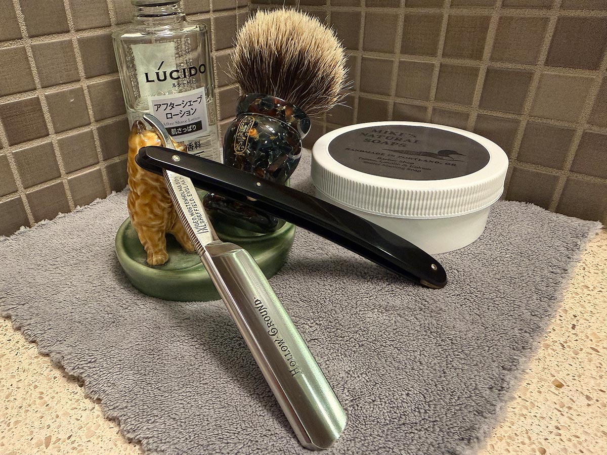 On a gray microfiber towel a straight razor with blade open about 45 degrees leans next to a bottle of aftershave and a jar of shave soap. A shave brush rests bristles-up in a green Wade ceramic pipe rest that features a brown Cairn Terrier.