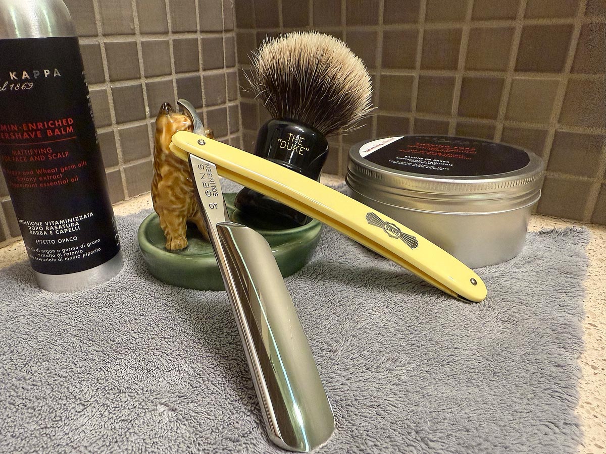 On a gray microfiber towel a straight razor with blade open about 45 degrees leans next to a bottle of aftershave and a jar of shave soap. A shave brush rests bristles-up in a green Wade ceramic pipe rest that features a brown Cairn Terrier.