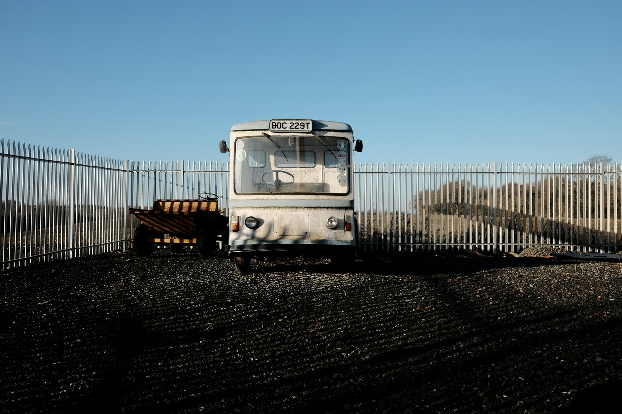 A dirty white electric milk float and a rusty yellow dumper truck parked side by side in a fenced gravel area.