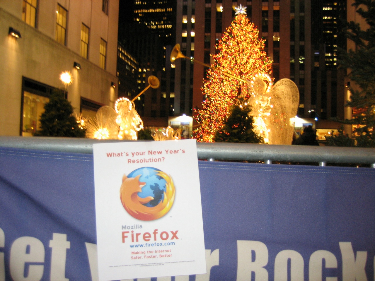 A flyer promoting Firefox hangs on a barricade near the Rockefeller Center Christmas tree in New York City. The flyer reads, "What's your New Year's resolution? / Mozilla Firefox / www.firefox.com / Making the Internet Safer, Faster, Better"