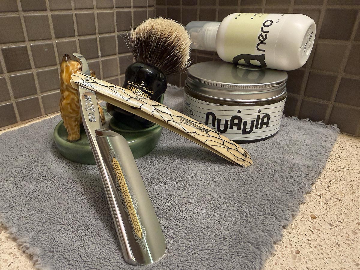 On a gray microfiber towel a straight razor with blade open about 45 degrees leans next to a bottle of aftershave and a jar of shave soap. A shave brush rests bristles-up in a green Wade ceramic pipe rest that features a brown Cairn Terrier.