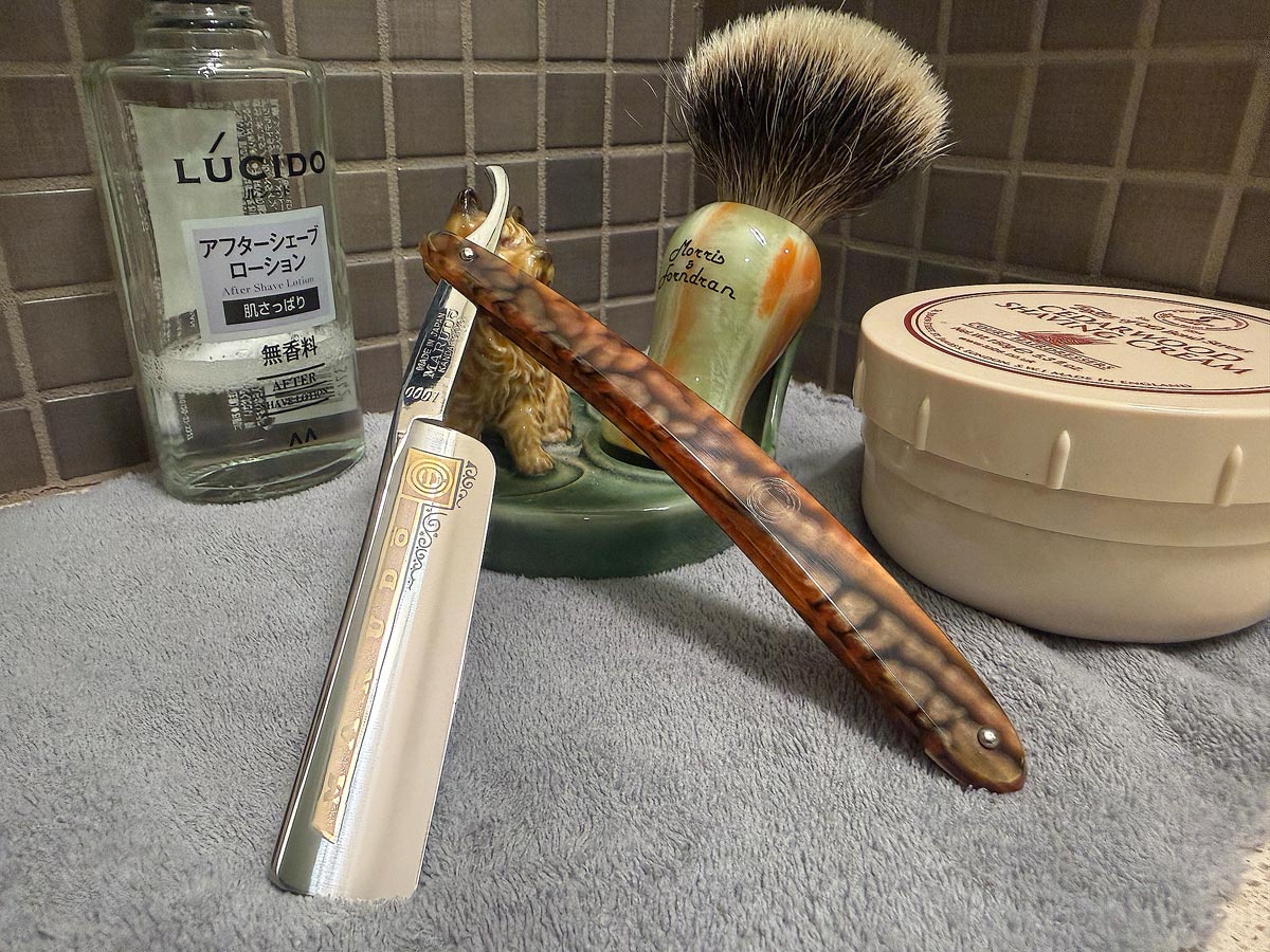On a gray microfiber towel a straight razor with blade open about 45 degrees leans next to a bottle of aftershave and a jar of shave soap. A shave brush rests bristles-up in a green Wade ceramic pipe rest that features a brown Cairn Terrier.