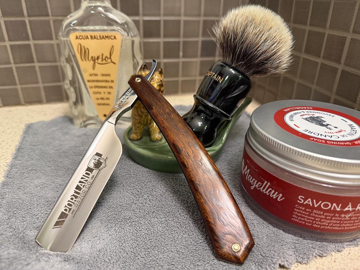 On a gray microfiber towel a straight razor with blade open about 45 degrees leans next to a bottle of aftershave and a jar of shave soap. A shave brush rests bristles-up in a green Wade ceramic pipe rest that features a brown Cairn Terrier.