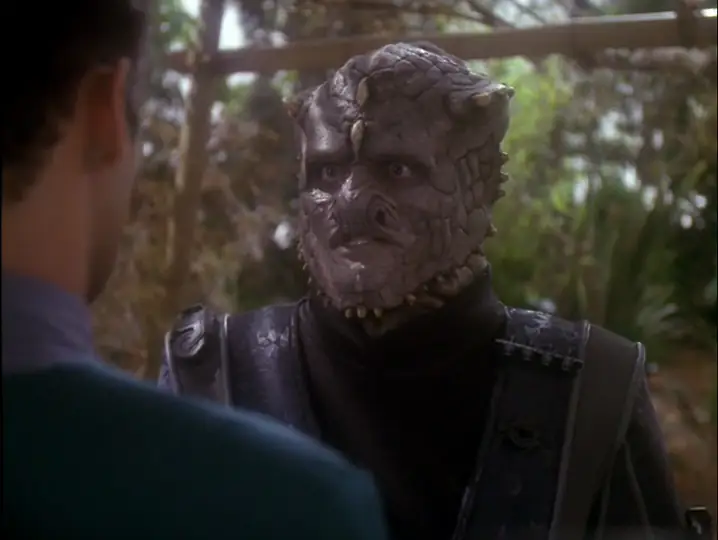 A close up shot of Scott MacDonald in Jem'Hadar makeup playing Goran'Agar. We're looking over the shoulder of Julian Bashir, who is wearing his teal colored uniform.