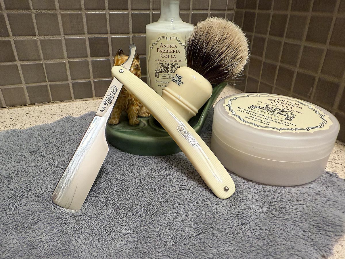 On a gray microfiber towel a straight razor with blade open about 45 degrees leans next to a bottle of aftershave and a jar of shave soap. A shave brush rests bristles-up in a green Wade ceramic pipe rest that features a brown Cairn Terrier.