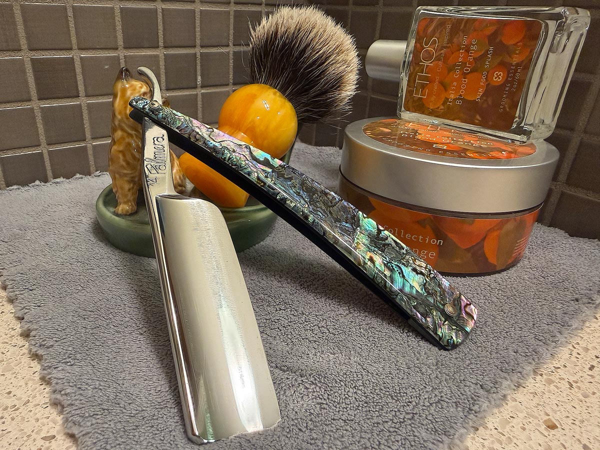 On a gray microfiber towel a straight razor with blade open about 45 degrees leans next to a bottle of aftershave and a jar of shave soap. A shave brush rests bristles-up in a green Wade ceramic pipe rest that features a brown Cairn Terrier.