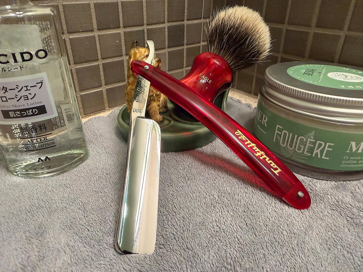 On a gray microfiber towel a straight razor with blade open about 45 degrees leans next to a bottle of aftershave and a jar of shave soap. A shave brush rests bristles-up in a green Wade ceramic pipe rest that features a brown Cairn Terrier.