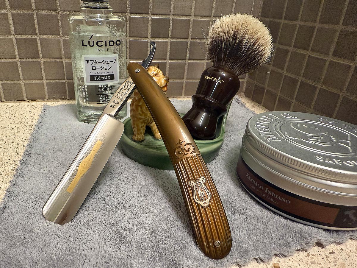 On a gray microfiber towel a straight razor with blade open about 45 degrees leans next to a bottle of aftershave and a jar of shave soap. A shave brush rests bristles-up in a green Wade ceramic pipe rest that features a brown Cairn Terrier.