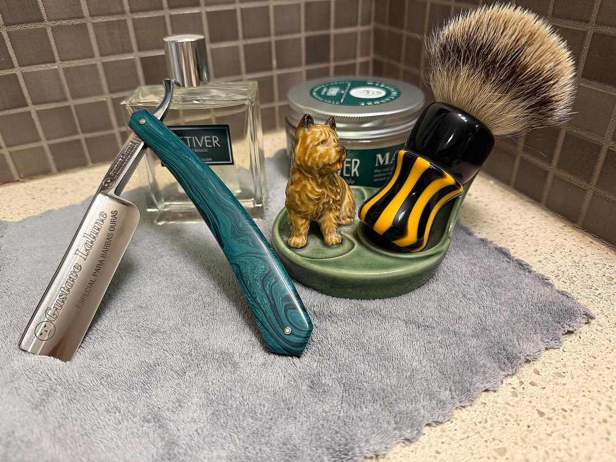 On a gray microfiber towel a straight razor with blade open about 45 degrees leans next to a bottle of aftershave and a jar of shave soap. A shave brush rests bristles-up in a green Wade ceramic pipe rest that features a brown Cairn Terrier.
