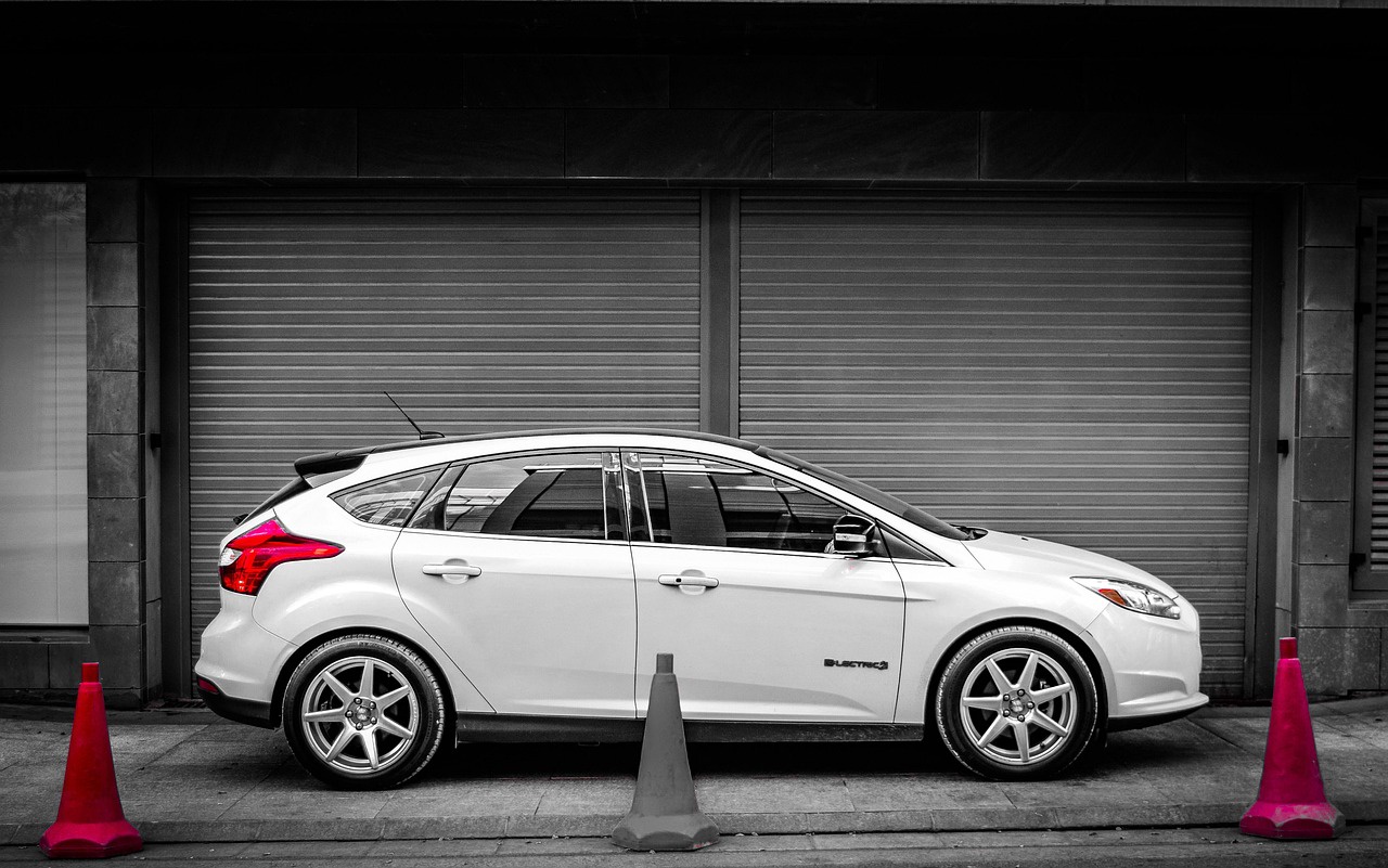 A white Ford Focus sits on a sidewalk (or pavement for U.K. readers) in front of two closed agarage doors. Three cones separate the street from the car.