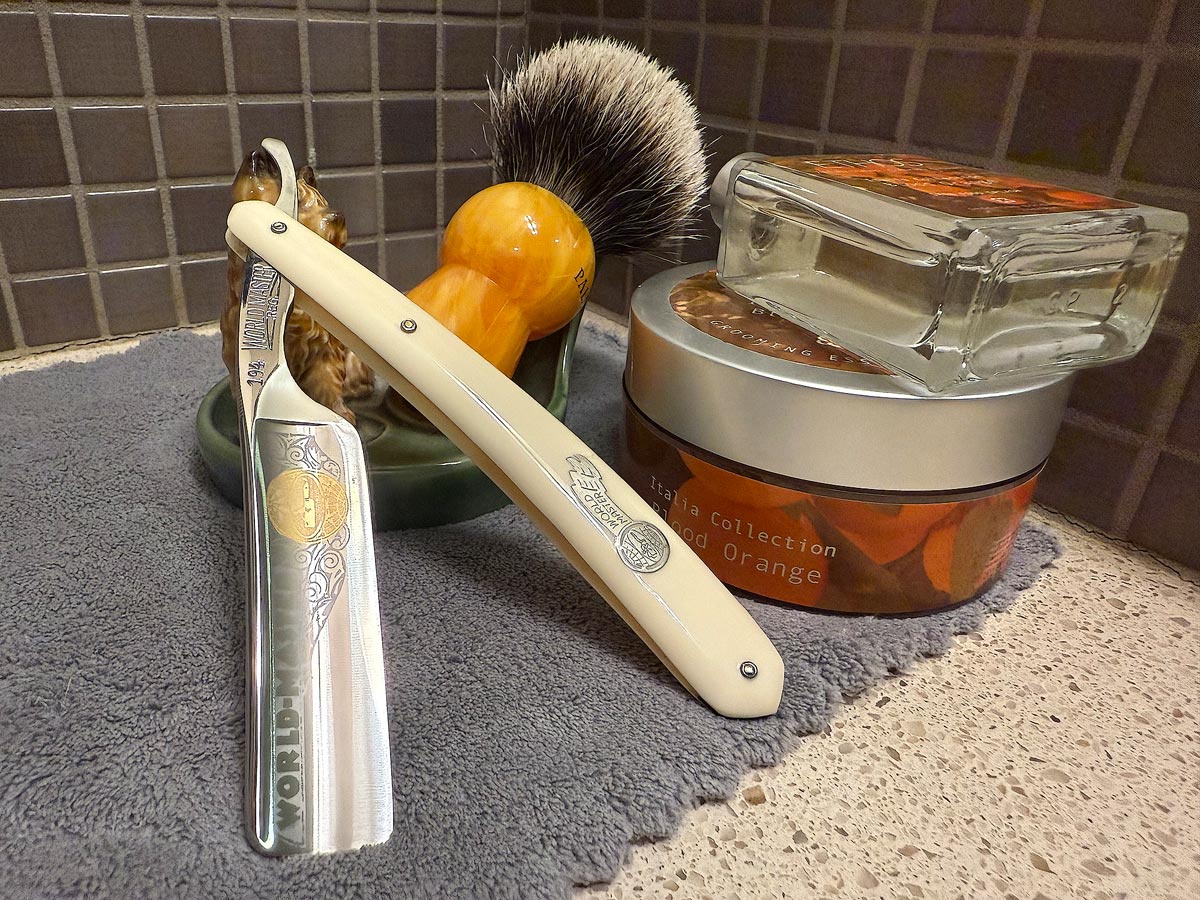 On a gray microfiber towel a straight razor with blade open about 45 degrees leans next to a bottle of aftershave and a jar of shave soap. A shave brush rests bristles-up in a green Wade ceramic pipe rest that features a brown Cairn Terrier.