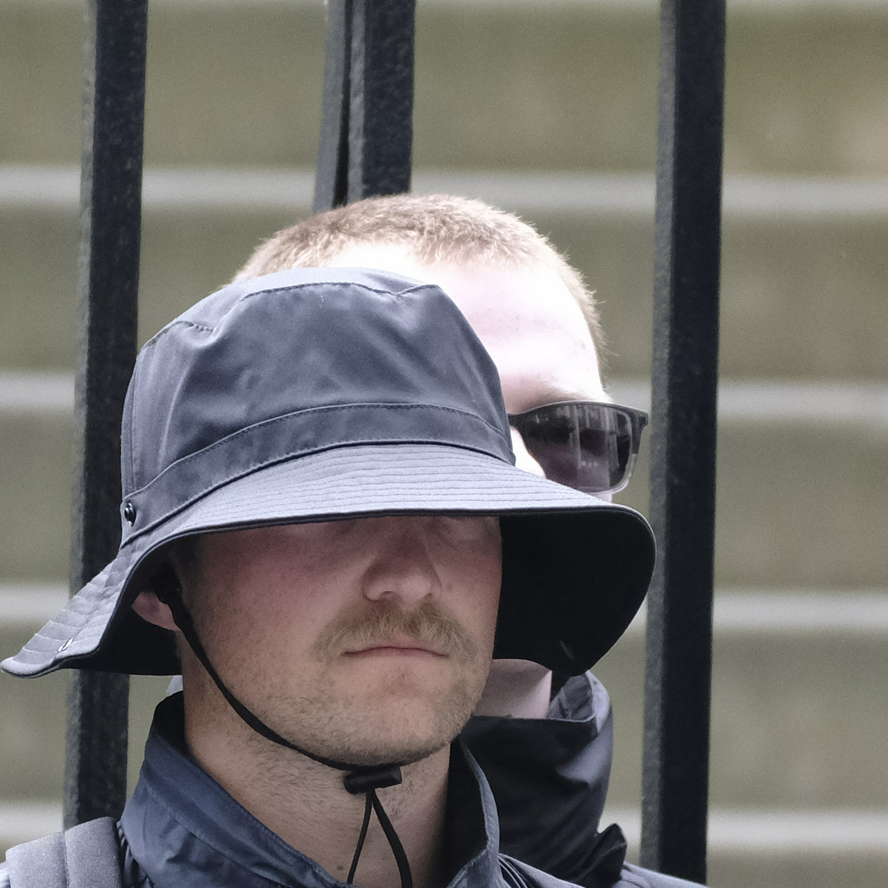 A pudgy guy with a dirty blonde buzzcut peers over the shoulder of the guy in front, with two-toned sunglasses