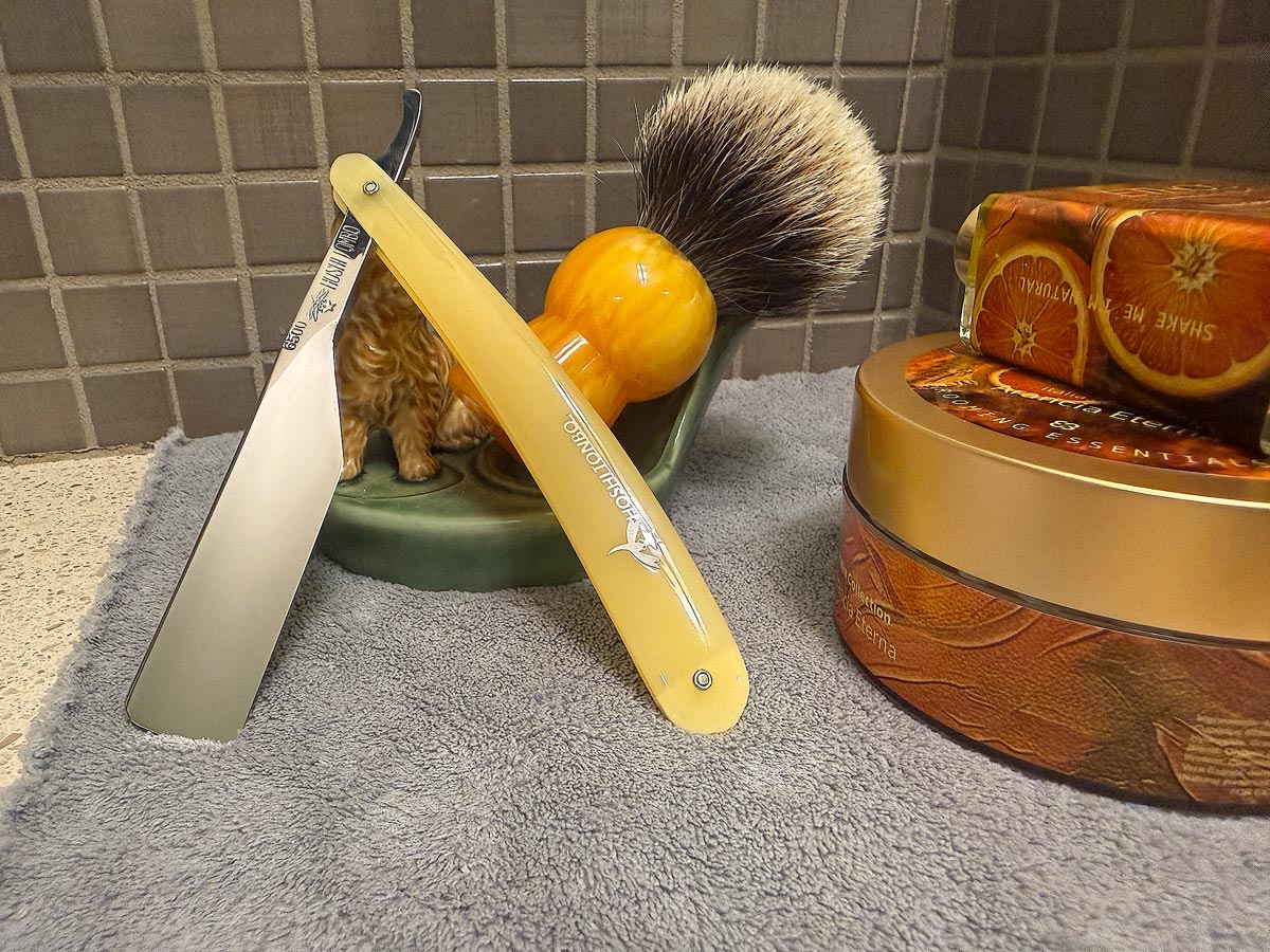 On a gray microfiber towel a straight razor with blade open about 45 degrees leans next to a bottle of aftershave and a jar of shave soap. A shave brush rests bristles-up in a green Wade ceramic pipe rest that features a brown Cairn Terrier.