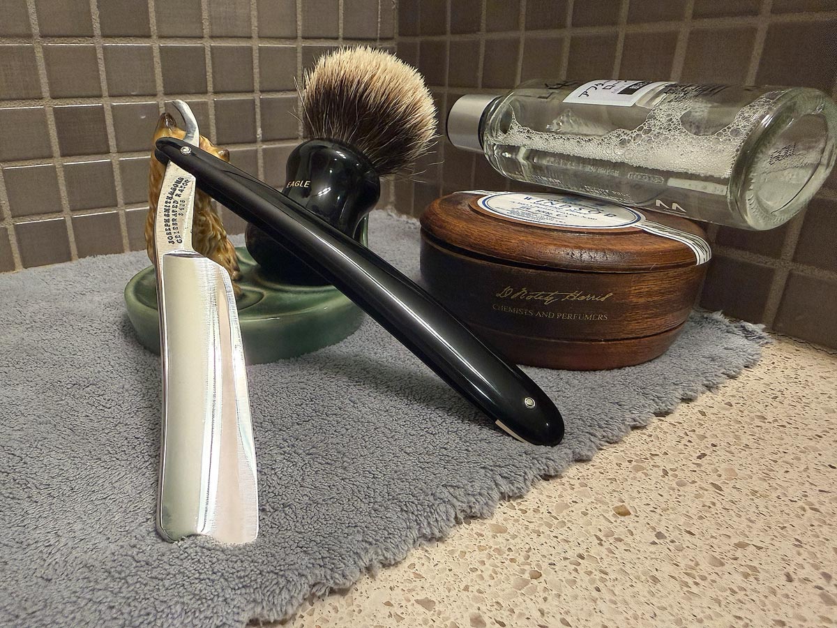 On a gray microfiber towel a straight razor with blade open about 45 degrees leans next to a bottle of aftershave and a jar of shave soap. A shave brush rests bristles-up in a green Wade ceramic pipe rest that features a brown Cairn Terrier.