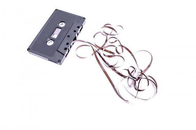 An audio cassette on a white surface. The cassette is opaque black and doesn't have a label on it. Several feet of tape have been pulled out and are coiled messily on thel same white surface the cassette rests on.