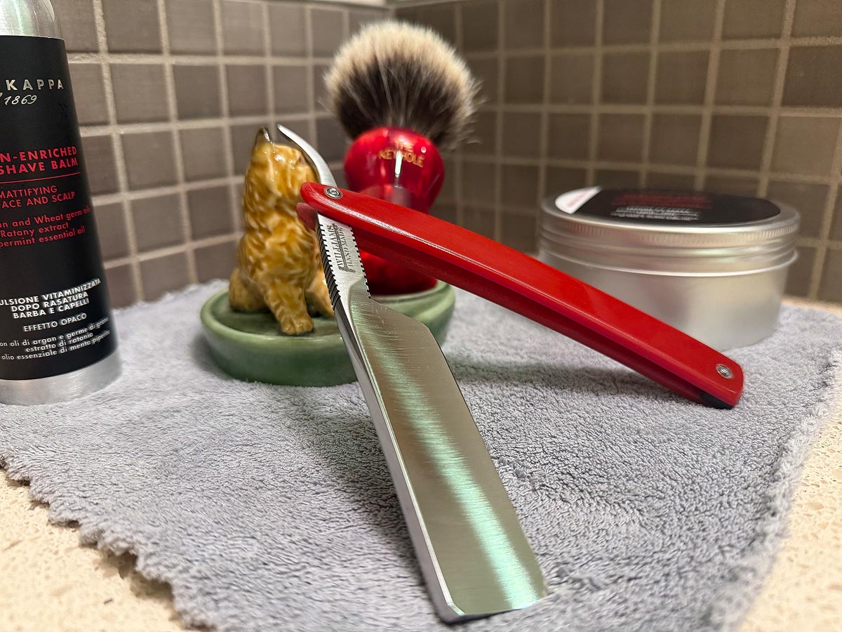 On a gray microfiber towel a straight razor with blade open about 45 degrees leans next to a bottle of aftershave and a jar of shave soap. A shave brush rests bristles-up in a green Wade ceramic pipe rest that features a brown Cairn Terrier.