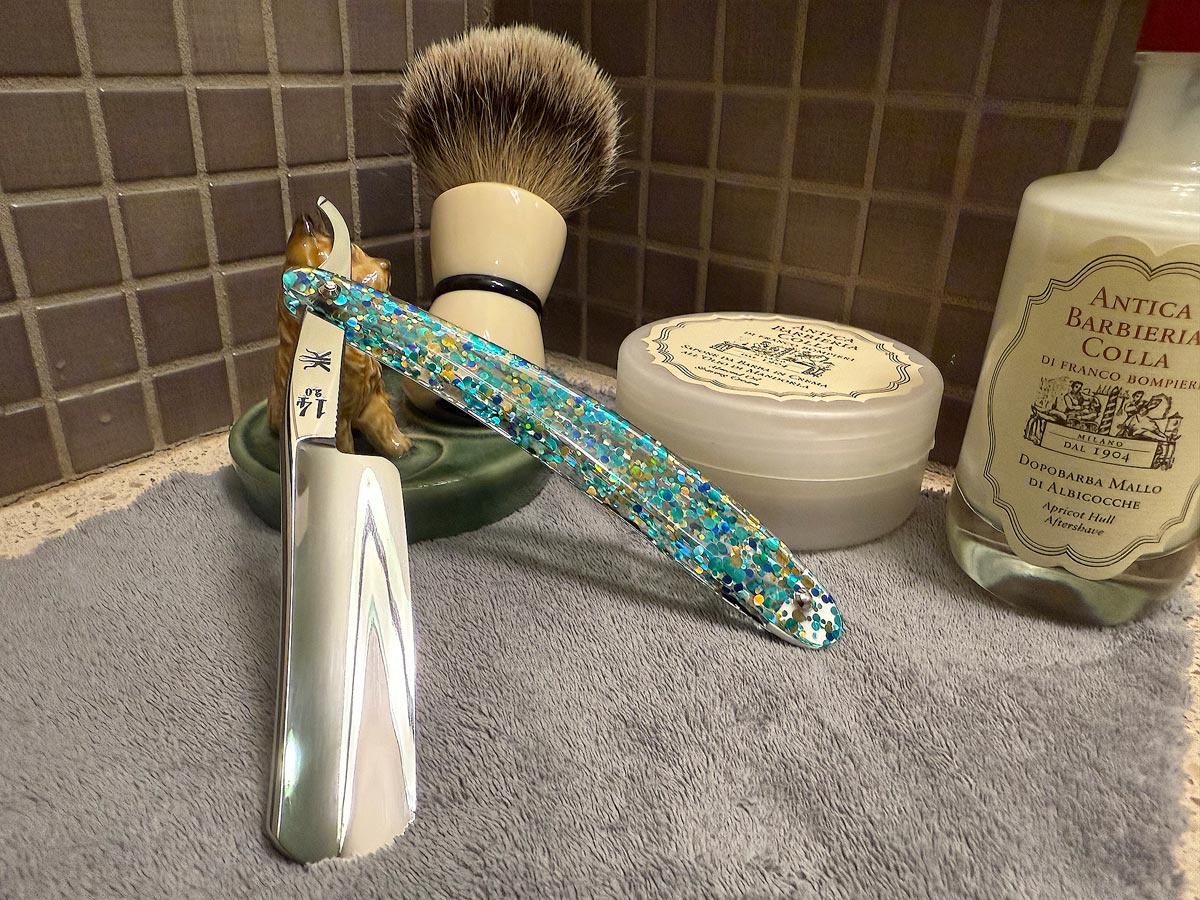 On a gray microfiber towel a straight razor with blade open about 45 degrees leans next to a bottle of aftershave and a jar of shave soap. A shave brush rests bristles-up in a green Wade ceramic pipe rest that features a brown Cairn Terrier.