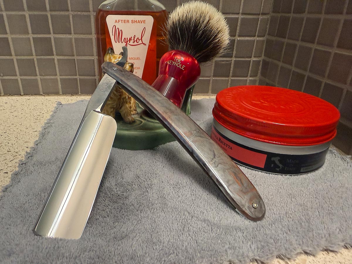 On a gray microfiber towel a straight razor with blade open about 45 degrees leans next to a bottle of aftershave and a jar of shave soap. A shave brush rests bristles-up in a green Wade ceramic pipe rest that features a brown Cairn Terrier.