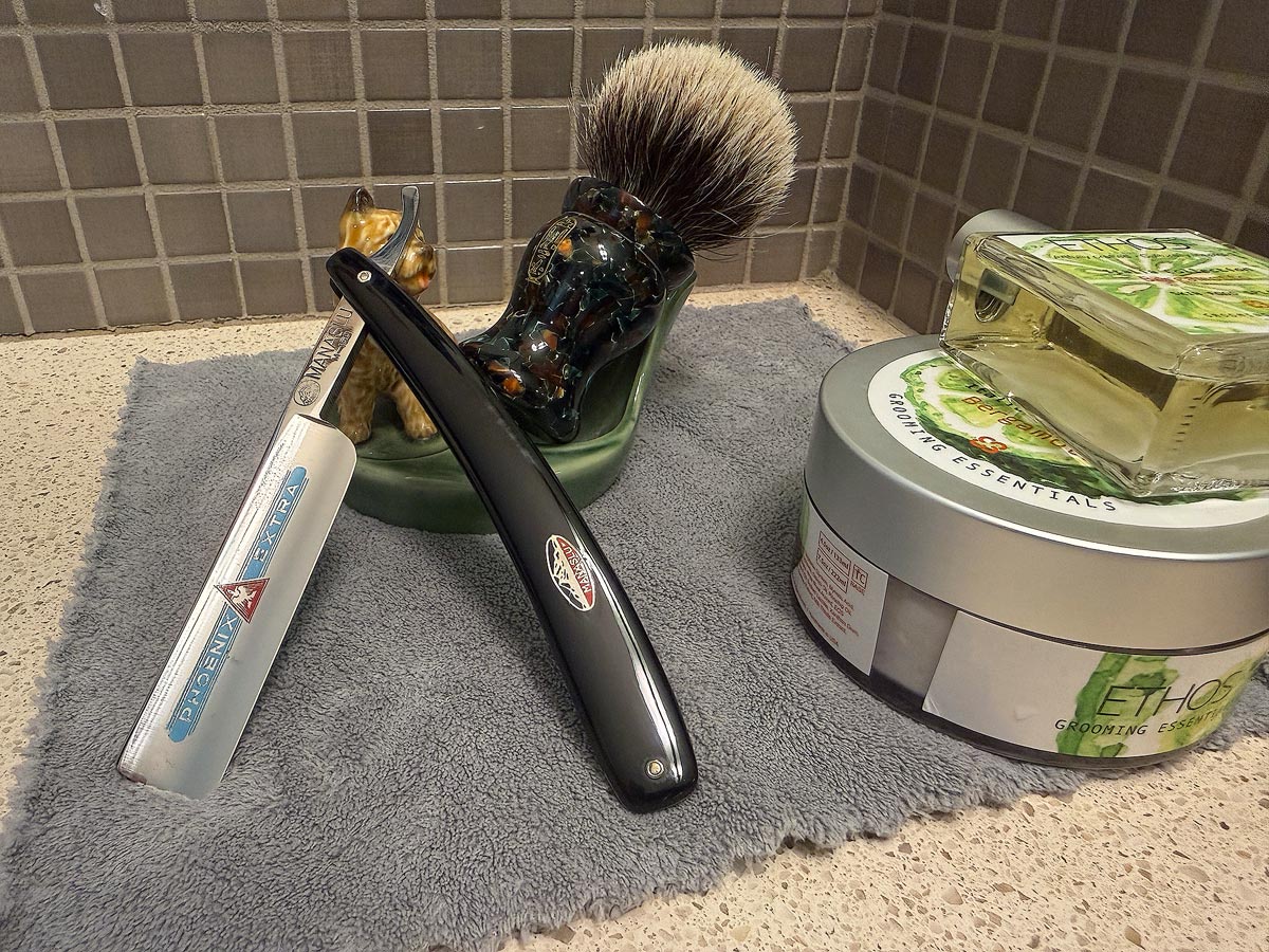On a gray microfiber towel a straight razor with blade open about 45 degrees leans next to a bottle of aftershave and a jar of shave soap. A shave brush rests bristles-up in a green Wade ceramic pipe rest that features a brown Cairn Terrier.