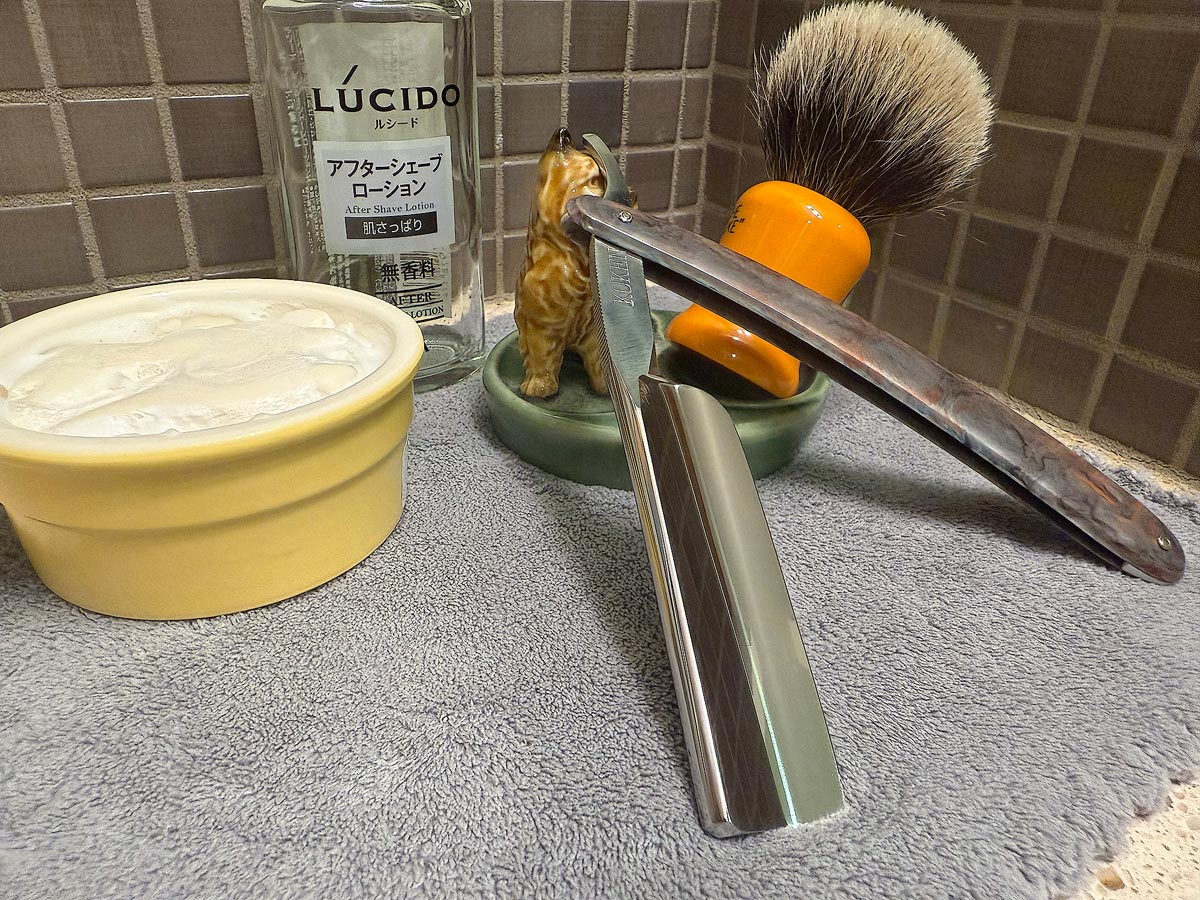 On a gray microfiber towel a straight razor with blade open about 45 degrees leans next to a bottle of aftershave and a jar of shave soap. A shave brush rests bristles-up in a green Wade ceramic pipe rest that features a brown Cairn Terrier.