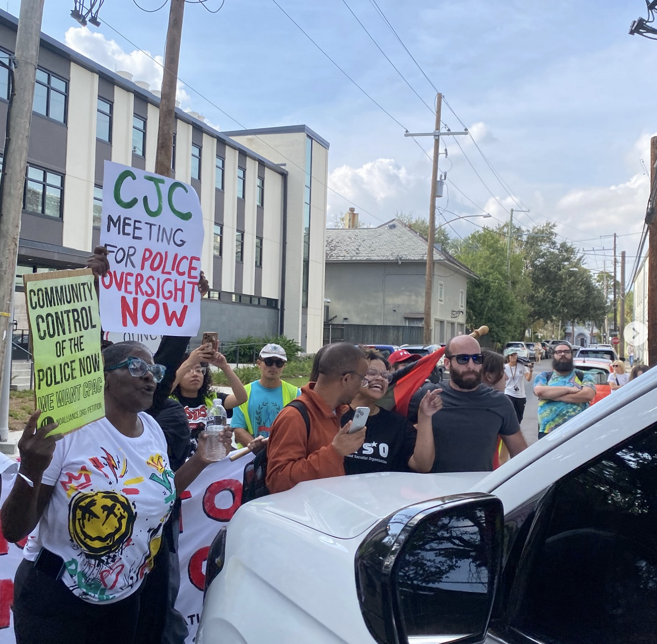 A crowd surrounds an NOPD vehicle as it tries to leave the court hearing, forcing it backwards down the street. Mothers Sean Coleman and Shanta Scott hold signs demanding police oversight as they fight for justice for their sons Junious Coleman and Jace Lee Scott, respectively.