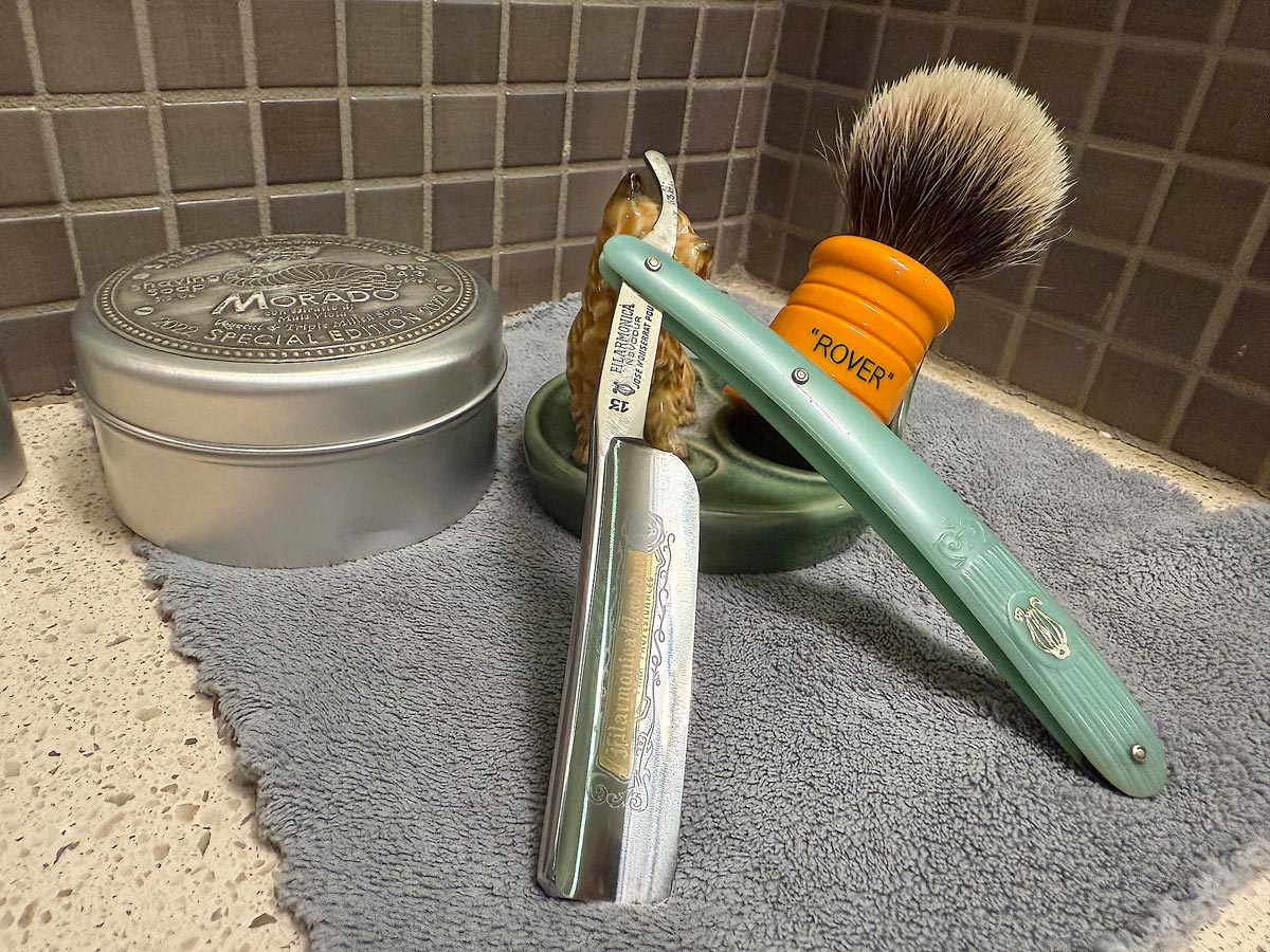 On a gray microfiber towel a straight razor with blade open about 45 degrees leans next to a bottle of aftershave and a jar of shave soap. A shave brush rests bristles-up in a green Wade ceramic pipe rest that features a brown Cairn Terrier.