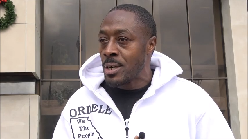 Charles Brown speaking on his case in 2021 in front of Cordele City Hall. | Getto Free Press NEWS G.B.R. A man wearing a hooded sweatshirt standing in front of a window.