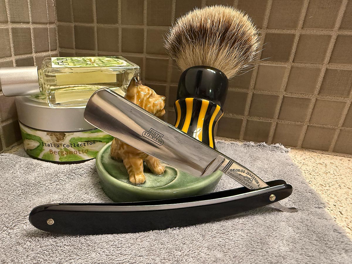 On a gray microfiber towel a straight razor with blade open about 45 degrees leans next to a bottle of aftershave and a jar of shave soap. A shave brush rests bristles-up in a green Wade ceramic pipe rest that features a brown Cairn Terrier.