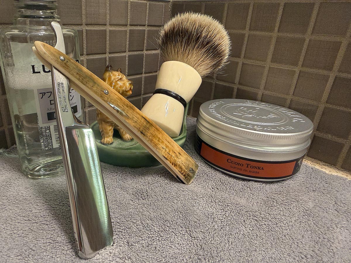 On a gray microfiber towel a straight razor with blade open about 45 degrees leans next to a bottle of aftershave and a jar of shave soap. A shave brush rests bristles-up in a green Wade ceramic pipe rest that features a brown Cairn Terrier.