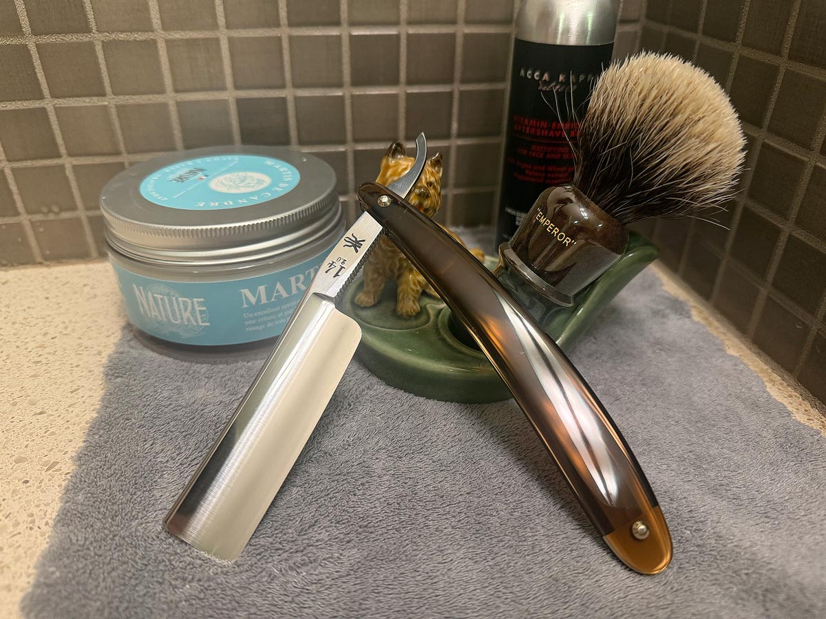 On a gray microfiber towel a straight razor with blade open about 45 degrees leans next to a bottle of aftershave and a jar of shave soap. A shave brush rests bristles-up in a green Wade ceramic pipe rest that features a brown Cairn Terrier.