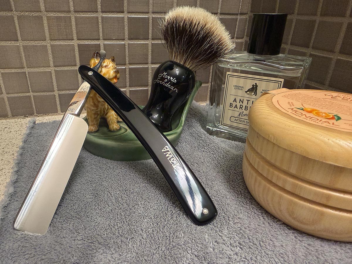 On a gray microfiber towel a straight razor with blade open about 45 degrees leans next to a bottle of aftershave and a jar of shave soap. A shave brush rests bristles-up in a green Wade ceramic pipe rest that features a brown Cairn Terrier.