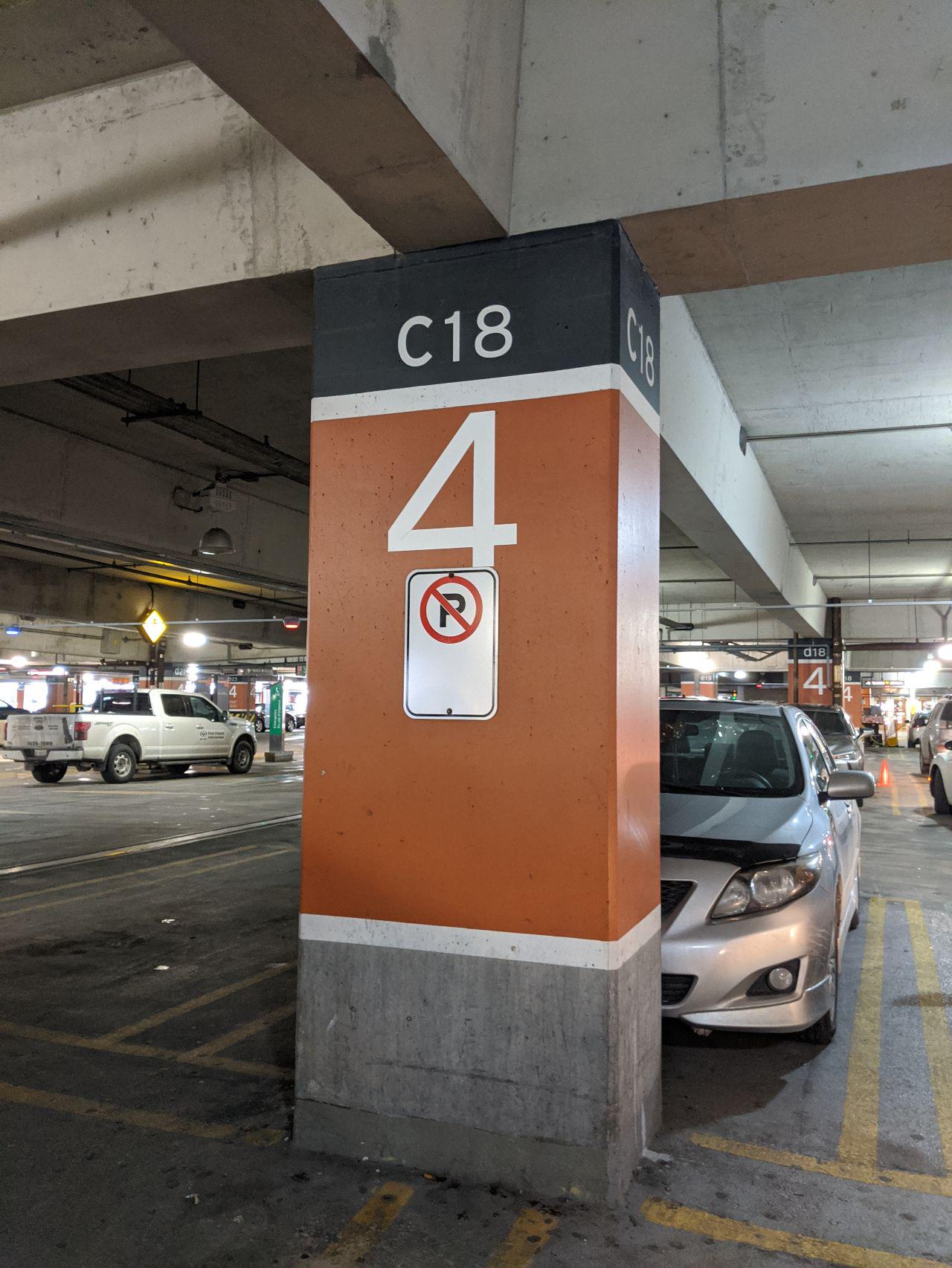 An orange pillar in an airport parking lot, it has a no parking sigh on one sided and has the test C18 / 4 painted onto it.