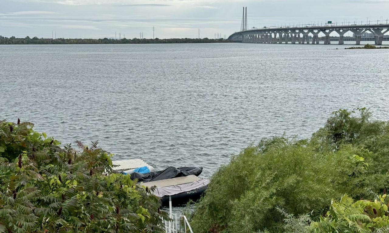 Vue sur le fleuve Saint-Laurent depuis la rive, avec le pont Samuel-de-Champlain à l'arrière-plan sous un ciel couvert.