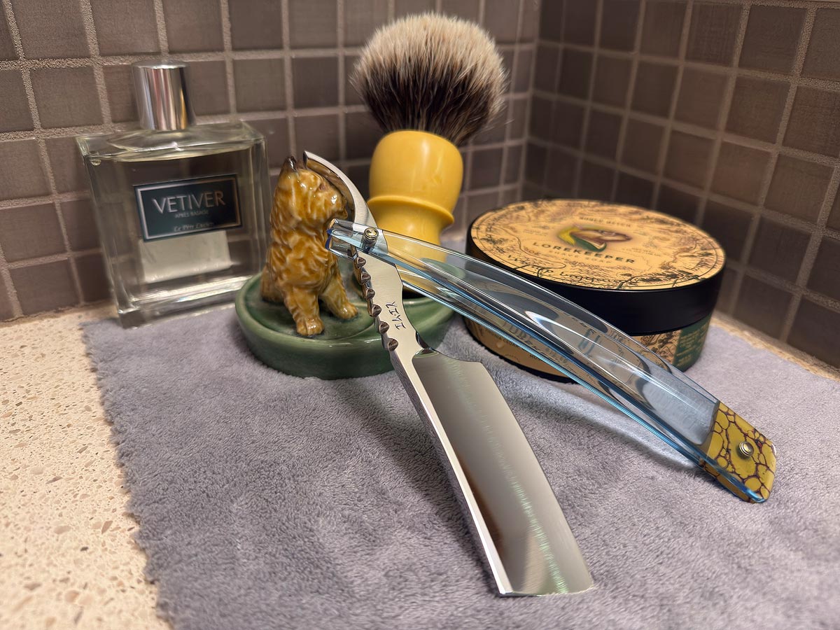 On a gray microfiber towel a straight razor with blade open about 45 degrees leans next to a bottle of aftershave and a jar of shave soap. A shave brush rests bristles-up in a green Wade ceramic pipe rest that features a brown Cairn Terrier.