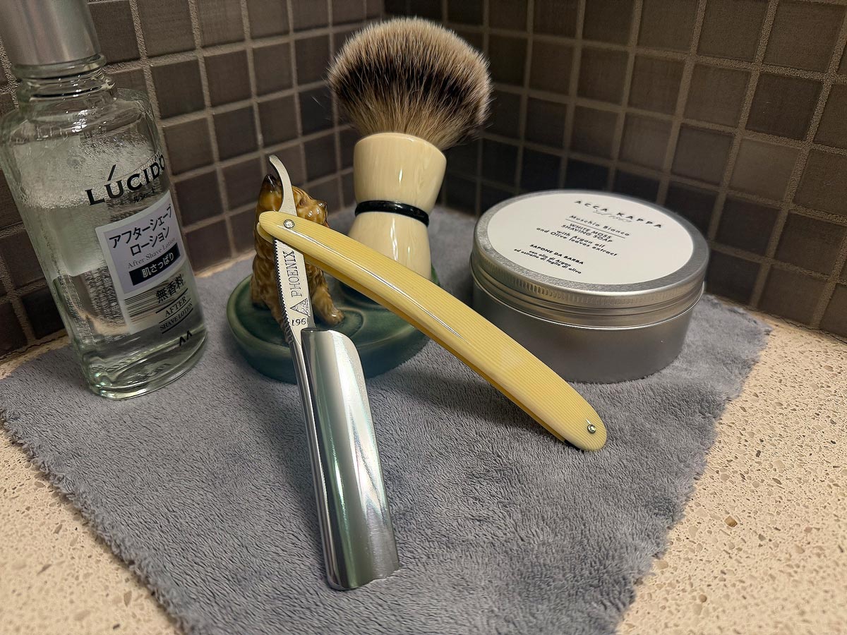 On a gray microfiber towel a straight razor with blade open about 45 degrees leans next to a bottle of aftershave and a jar of shave soap. A shave brush rests bristles-up in a green Wade ceramic pipe rest that features a brown Cairn Terrier.