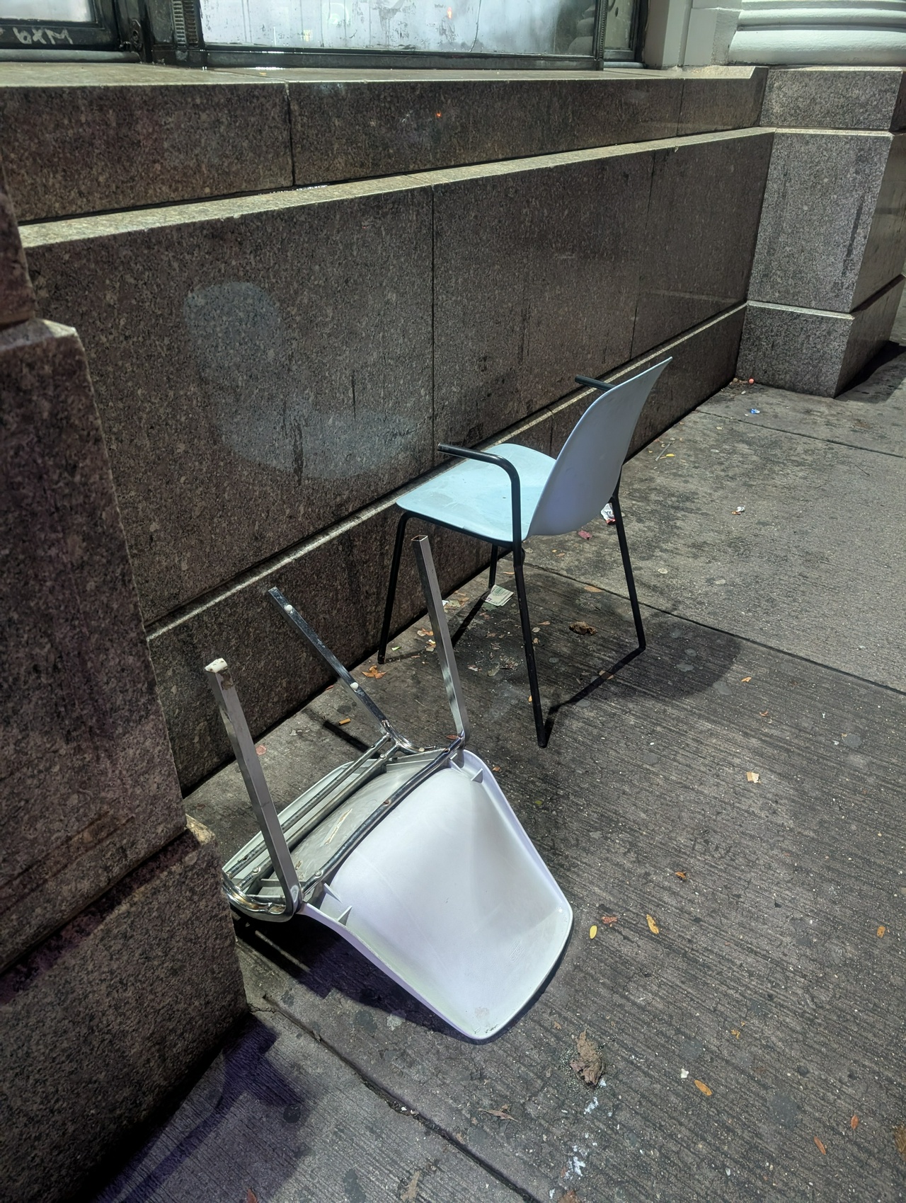 two plastic chairs are near a building wall in brooklyn. one has chonky metal legs, and is face down on the sidewalk, facing the street. the other has black legs and arms, and stares at the smooth stone wall, as if to look at its own reflection.