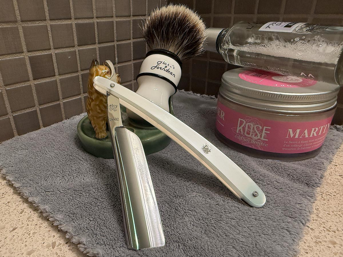 On a gray microfiber towel a straight razor with blade open about 45 degrees leans next to a bottle of aftershave and a jar of shave soap. A shave brush rests bristles-up in a green Wade ceramic pipe rest that features a brown Cairn Terrier.