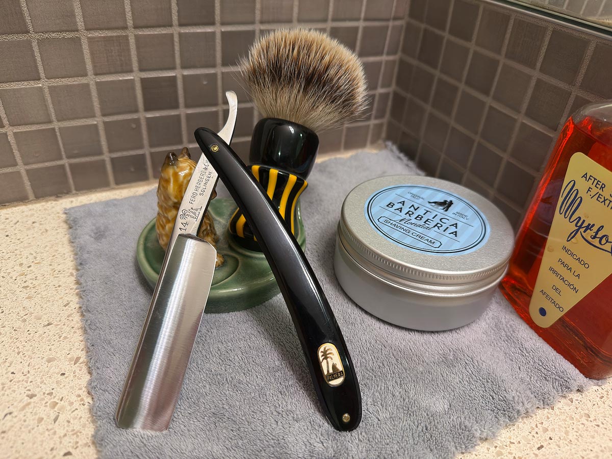 On a gray microfiber towel a straight razor with blade open about 45 degrees leans next to a bottle of aftershave and a jar of shave soap. A shave brush rests bristles-up in a green Wade ceramic pipe rest that features a brown Cairn Terrier.