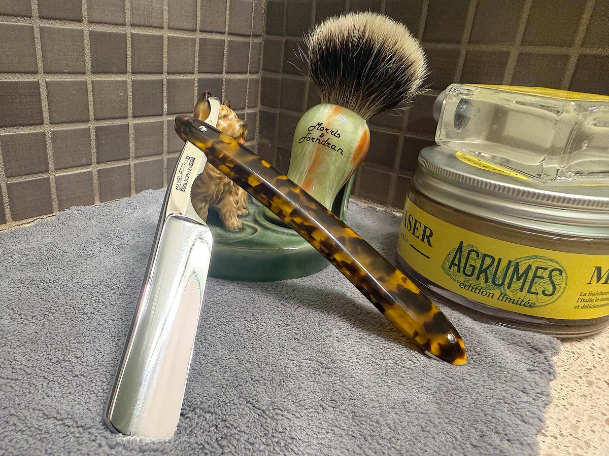 On a gray microfiber towel a straight razor with blade open about 45 degrees leans next to a bottle of aftershave and a jar of shave soap. A shave brush rests bristles-up in a green Wade ceramic pipe rest that features a brown Cairn Terrier.