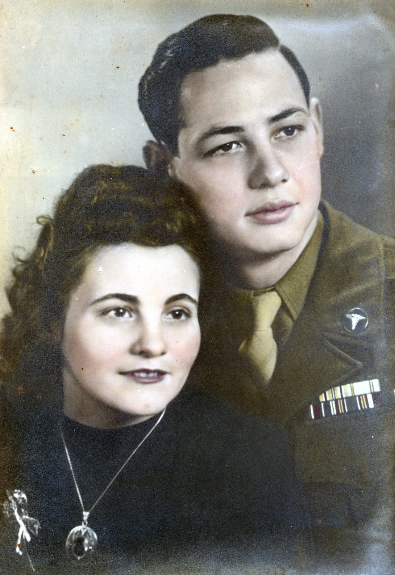 A vintage colorized portrait of a young couple; the woman is wearing a dark blouse and a necklace, resting her head on the shoulder of a man who is dressed in a military uniform with a beret and decorated with multiple ribbons on his chest. Both are looking directly at the camera, conveying a serene and affectionate demeanor.